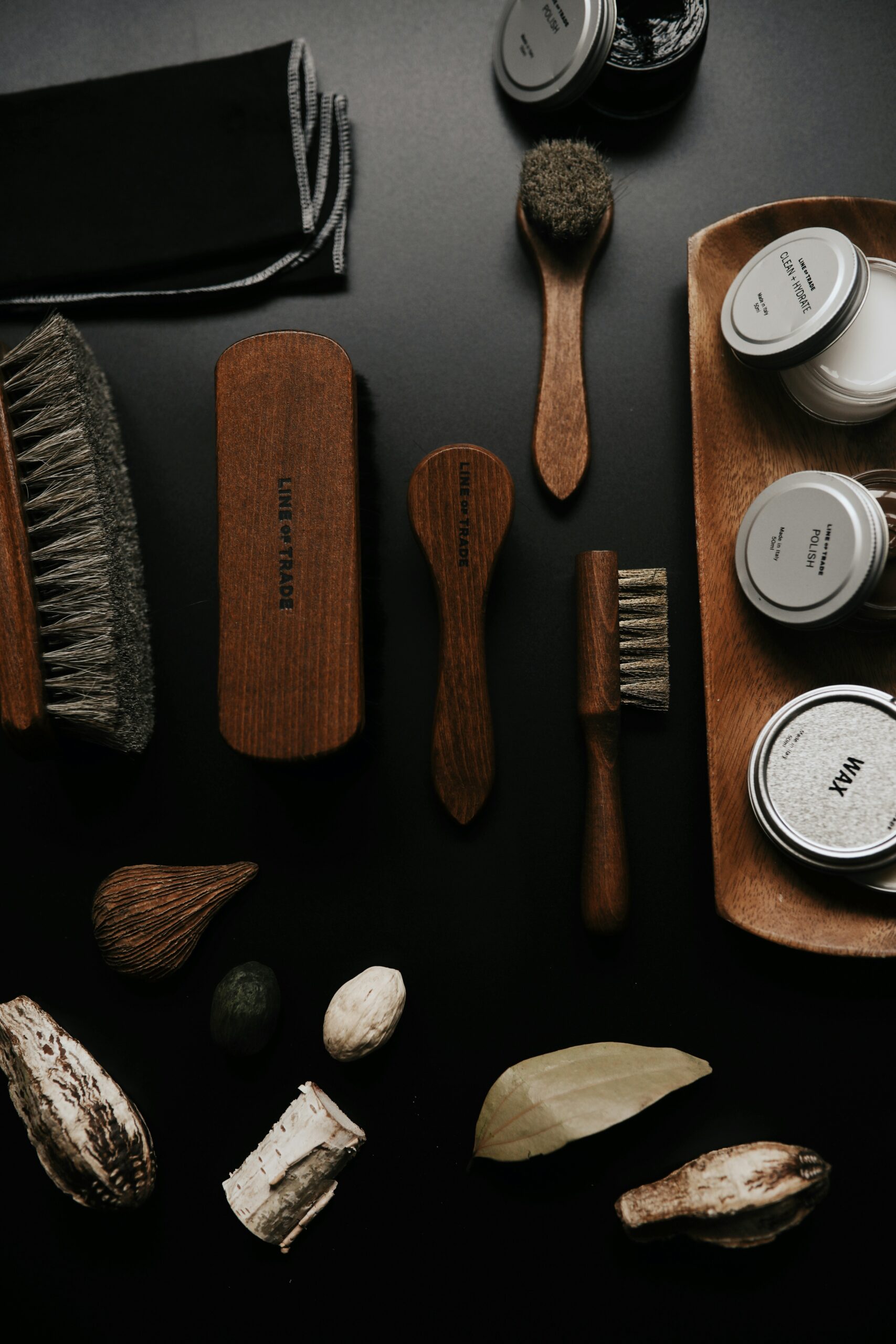 Flat lay of shoe care accessories including brushes, tins of wax and polish, cloth, and wooden tray arranged neatly on a black surface